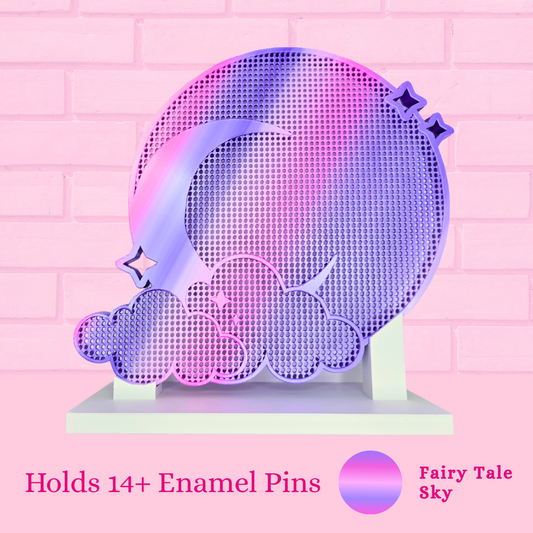A decorative acrylic pin holder shaped like a crescent moon and clouds, shown on a pink brick wall background. The holder has a holographic purple-pink-blue gradient finish and includes text at the bottom reading 'Holds 14+ Enamel Pins' and 'Fairy Tale Sky' with a matching color swatch.