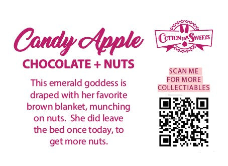 The back of the character card reads: Candy Apple Chocolate + Nuts, This emerald goddess is draped with her favorite brown blanket, munching on nuts. She did leave the bed once today, to get more nuts.
