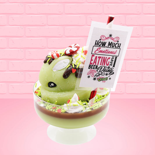 Cute kawaii ice cream Emotional Grinch Fudge sundae which has a sign and green white and red heart sprinkles inside of the dish as well as swirls of whipped cream. The bunny has chocolate glaze, peppermint candies and a swirl of whipped cream on it’s head. The sign says How much emotional eating have I been doing?