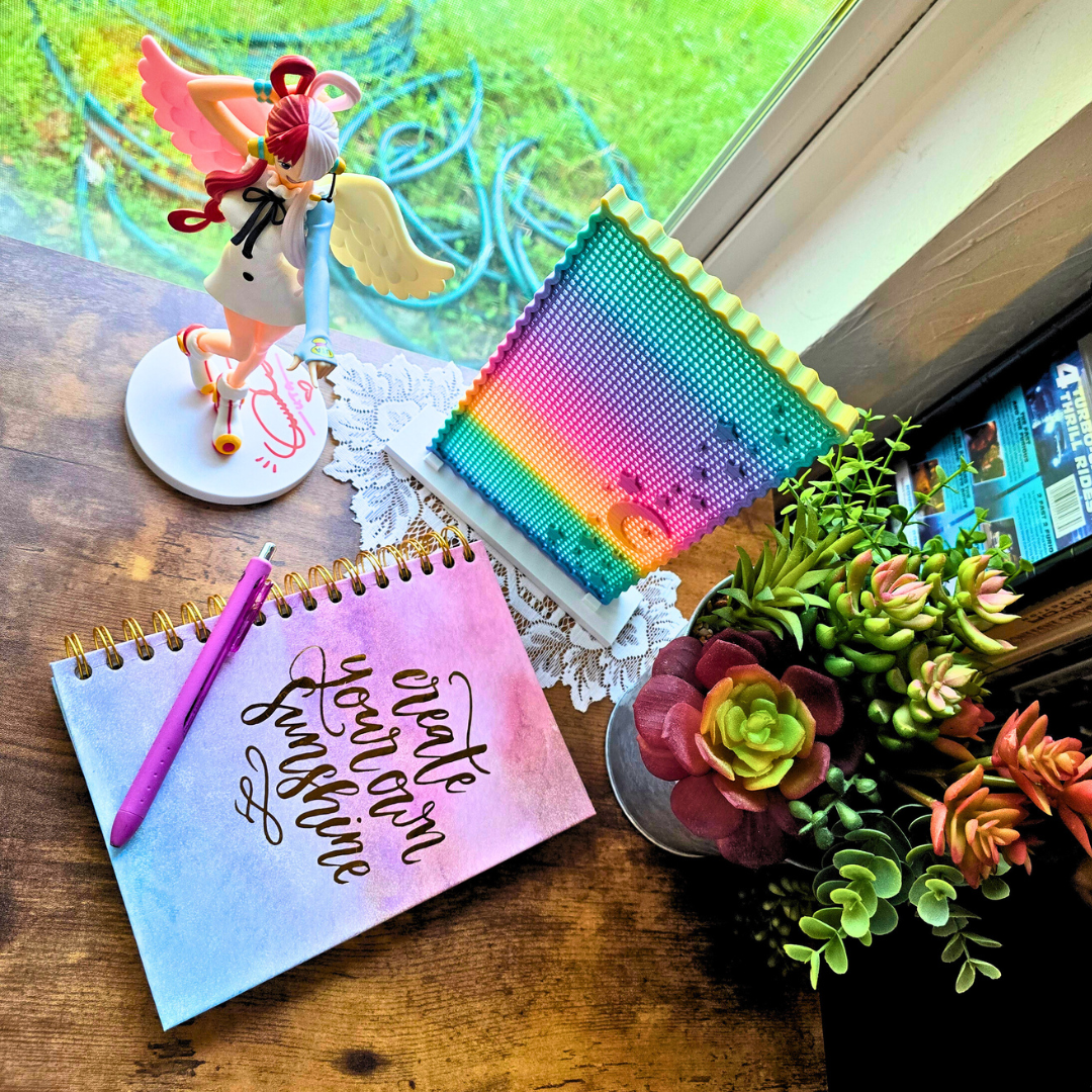 Example display of Moon and Star enamel pin board with an anime girl figure on a desk next to plants and a journal that says create your own sunshine at a different angle.