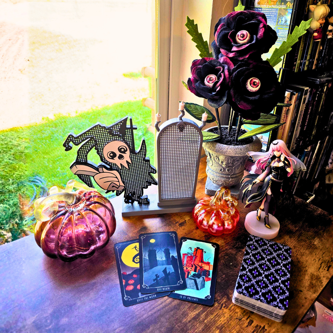L-Death with Mirror enamel pin board next to two glass pumpkins, tarot deck, eyeball flowers and a goth anime girl figure on top of a desk in a room.