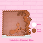 A decorative wooden pin holder with a hand-crafted bird and floral design, shown on a white stand against a pink brick wall background. The image includes color swatches labeled 'White' and 'Bread Brown' and text at the bottom stating 'Holds 14+ Enamel Pins'.
