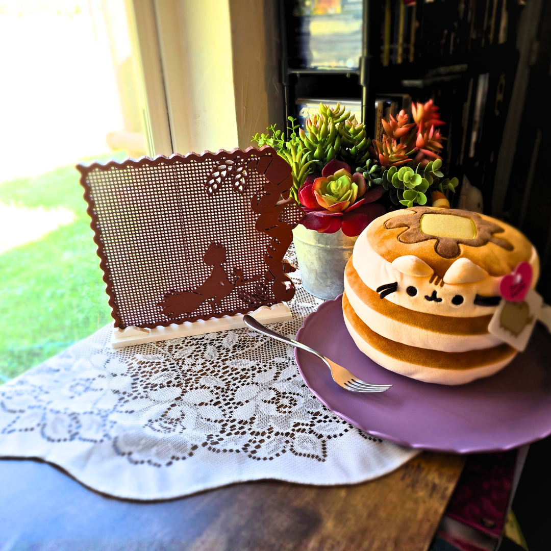 Bread Brown Kiki’s Bread Bakery enamel pin display on top of a doily, next to a cute cat pancake plush on a purple plate.