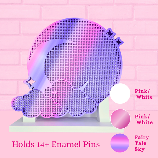 A pink and purple holographic acrylic stand shaped like a crescent moon and clouds, displayed against a pink brick wall. The text at the bottom reads 'Holds 14+ Enamel Pins' with color swatches labeled 'Pink/White' and 'Fairy Tale Sky'.