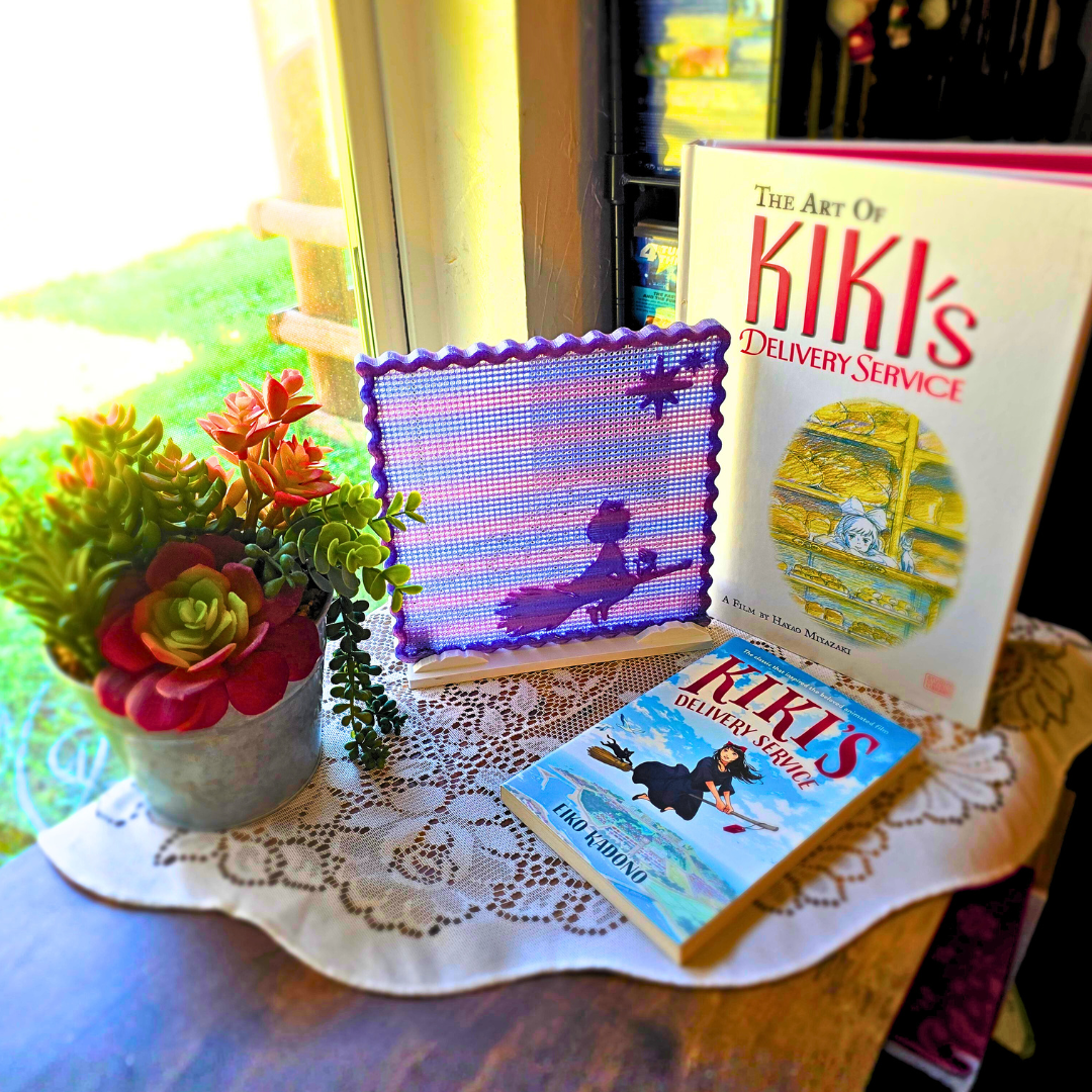 Example display of Kiki's Skyenamel pin board next to books on a desk next to plant on a large doily by a window in a room.