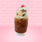 Cute kawaii bunny Root Beer Float white bunny in a brown drink with a black and white straw on the side. The bunny has a strawberry, brown glitter, silver pearls, on top next to the swirl of whipped cream. This view is from the side at an angle.