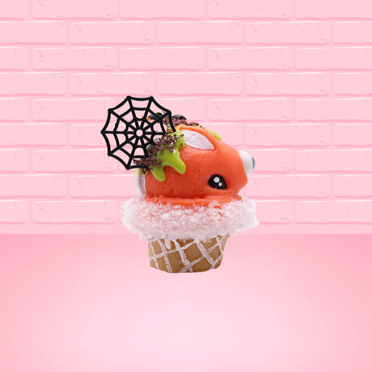 Cute kawaii bunny Monster Scoop orange ice cream cone with a blue eye in the center, green drizzle with chocolate bits, pumpkin sprinkles, a black spider and web near the top of the bunny next to swirl of the whipped cream. This view is from the side.