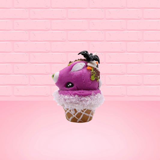 Cute kawaii bunny Monster Scoop purple ice cream cone with a green eye in the center, green drizzle with chocolate bits, pumpkin and bat sprinkles, and a bat on the top of the bunny near the whipped cream swirl. This view is from the side.