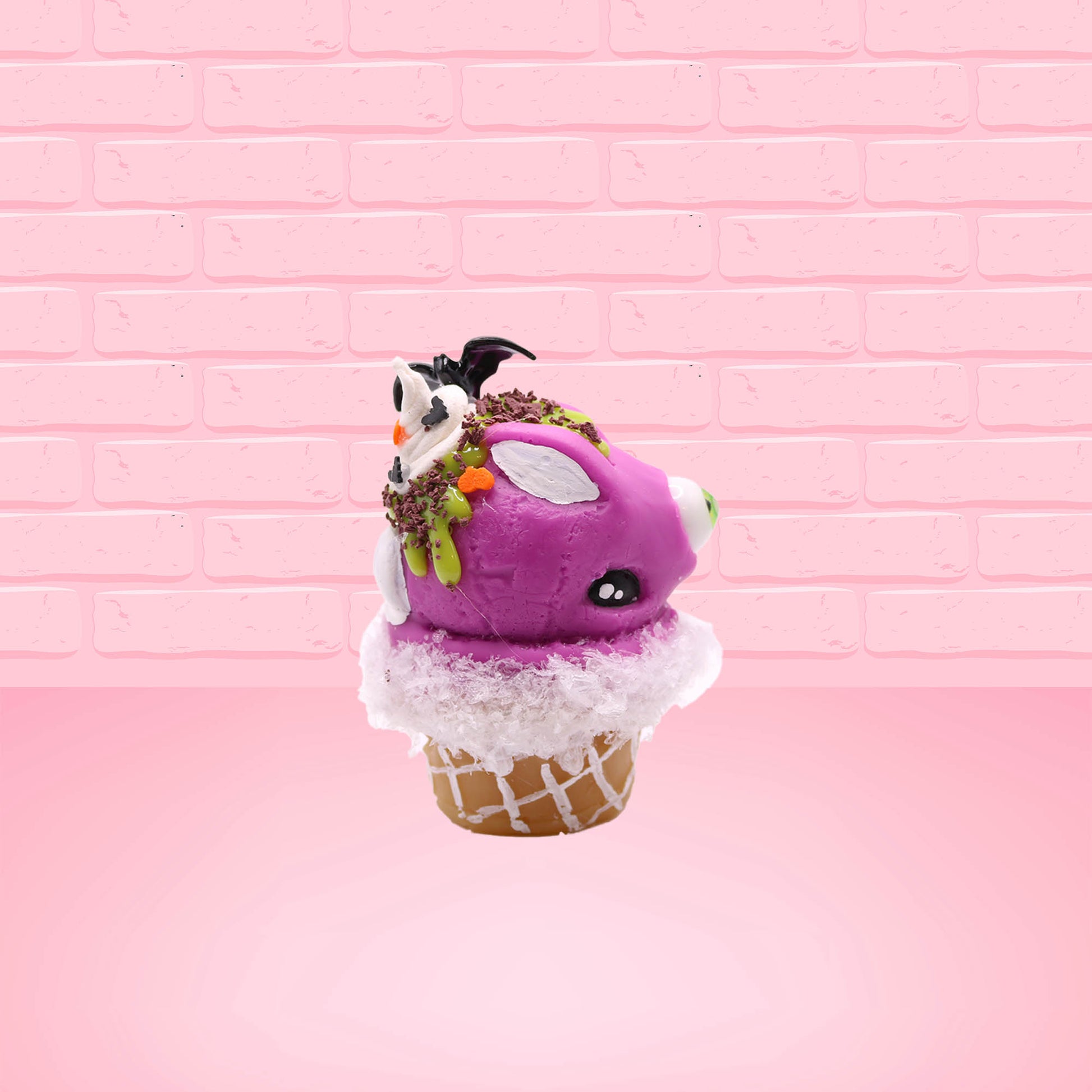 Cute kawaii bunny Monster Scoop purple ice cream cone with a green eye in the center, green drizzle with chocolate bits, pumpkin and bat sprinkles, and a bat on the top of the bunny near the whipped cream swirl. This view is from the other side.