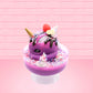 Cute kawaii unicorn bunny Coco Dazzle Star purple ice cream with pink, purple and white sprinkles with swirls of whipped cream in the dish. The unicorn bunny has a strawberry and banana slices with chocolate drizzle and sprinkles near the whipped cream swirl. This view is from the side.