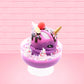 Cute kawaii unicorn bunny Coco Dazzle Star purple ice cream with pink, purple and white sprinkles with swirls of whipped cream in the dish. The unicorn bunny has a strawberry and banana slices with chocolate drizzle and sprinkles near the whipped cream swirl. This view is from the other side.