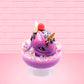 Cute kawaii unicorn bunny Coco Dazzle Star purple ice cream with pink, purple and white sprinkles with swirls of whipped cream in the dish. The unicorn bunny has a strawberry and banana slices with chocolate drizzle and sprinkles near the whipped cream swirl. This view is from the front at an angle.