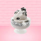 Cute kawaii bunny cookies and cream ice cream white scoop sundae with whipped cream swirls and black white crumbles in the dish. A black and white sandwhich cookie is on top of the resin bunny, sprinkled with black and white crumbles and a swirl of whipped cream. This view is from the side.