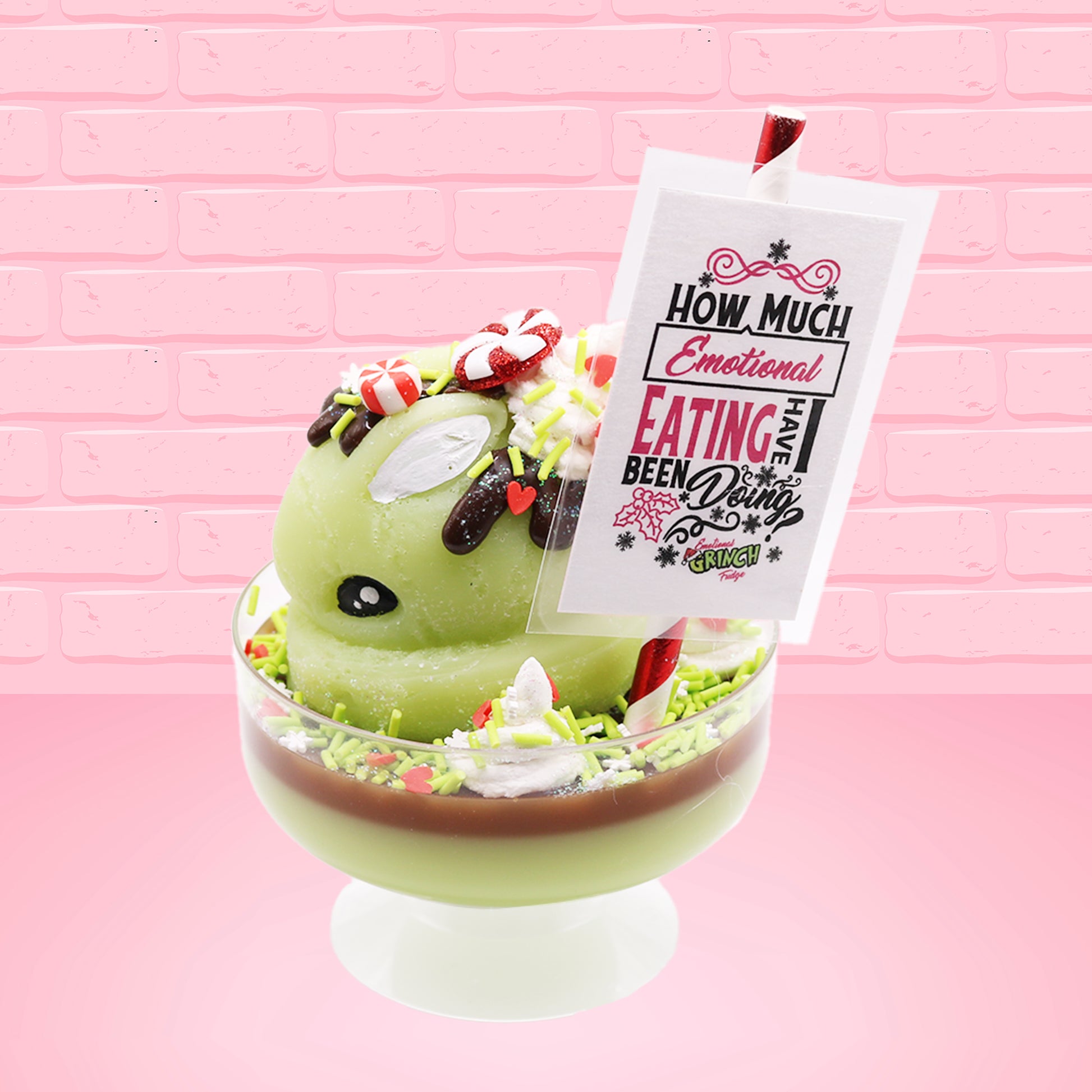 Cute kawaii ice cream Emotional Grinch Fudge sundae which has a sign and green white and red heart sprinkles inside of the dish as well as swirls of whipped cream. The bunny has chocolate glaze, peppermint candies and a swirl of whipped cream on it’s head. The sign says How much emotional eating have I been doing?