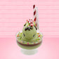 Cute kawaii ice cream Emotional Grinch Fudge sundae which has a sign and green white and red heart sprinkles inside of the dish as well as swirls of whipped cream. The bunny has chocolate glaze, peppermint candies and a swirl of whipped cream on it’s head. This view is from the front and shows the pole of the sign.