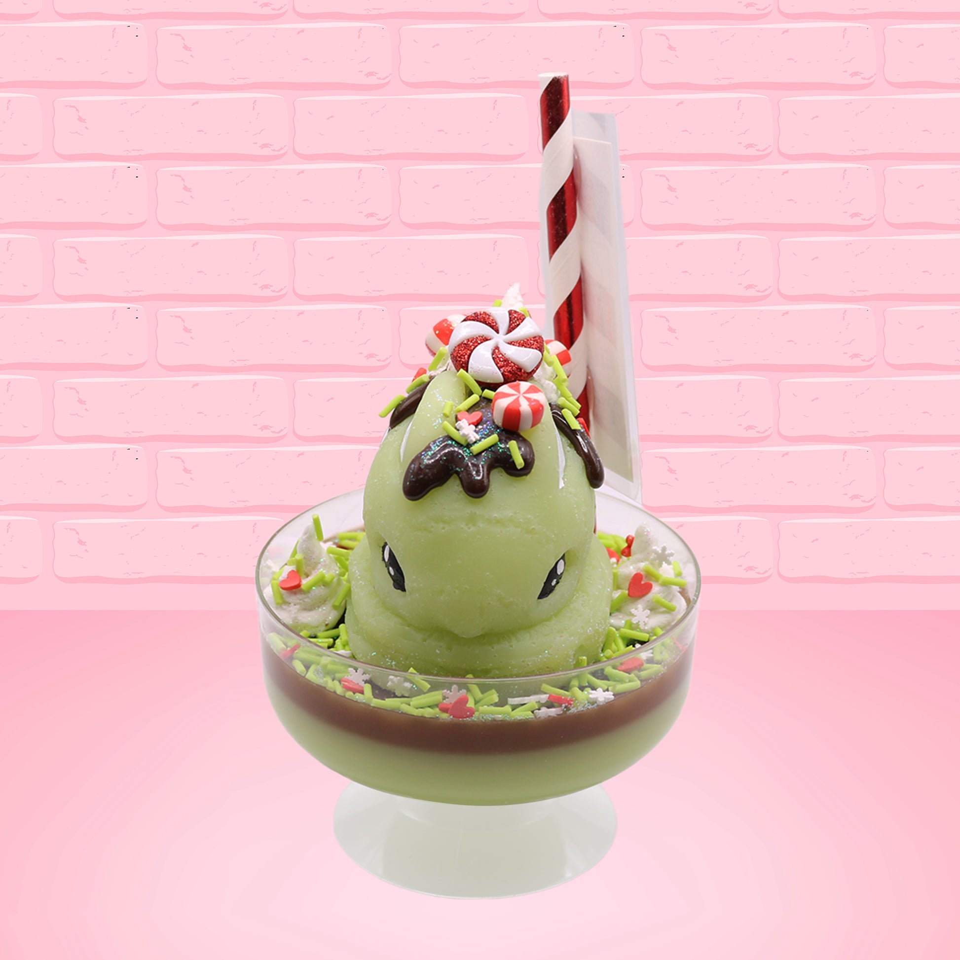 Cute kawaii ice cream Emotional Grinch Fudge sundae which has a sign and green white and red heart sprinkles inside of the dish as well as swirls of whipped cream. The bunny has chocolate glaze, peppermint candies and a swirl of whipped cream on it’s head. This view is from the front and shows the pole of the sign.