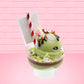 Cute kawaii ice cream Emotional Grinch Fudge sundae which has a sign and green white and red heart sprinkles inside of the dish as well as swirls of whipped cream. The bunny has chocolate glaze, peppermint candies and a swirl of whipped cream on it’s head. This view is from the side.