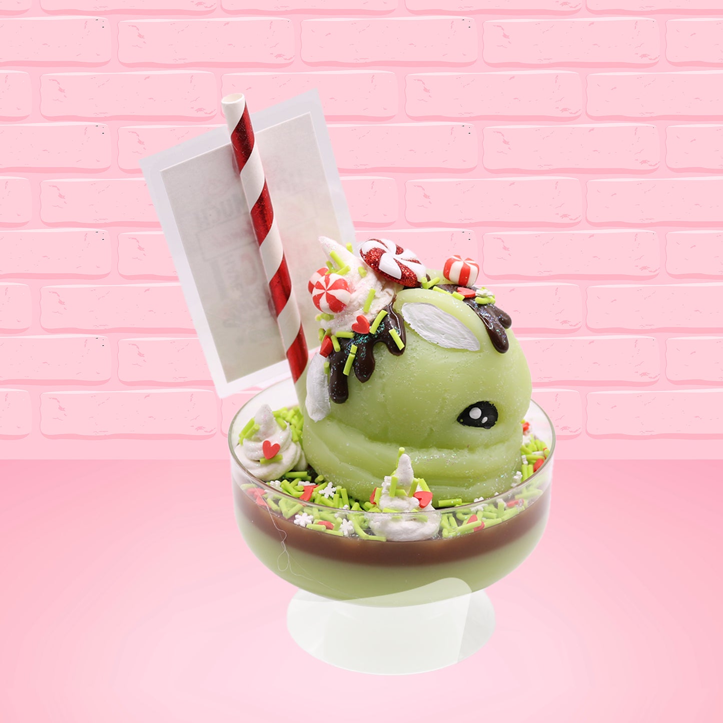 Cute kawaii ice cream Emotional Grinch Fudge sundae which has a sign and green white and red heart sprinkles inside of the dish as well as swirls of whipped cream. The bunny has chocolate glaze, peppermint candies and a swirl of whipped cream on it’s head. This view is from the side.