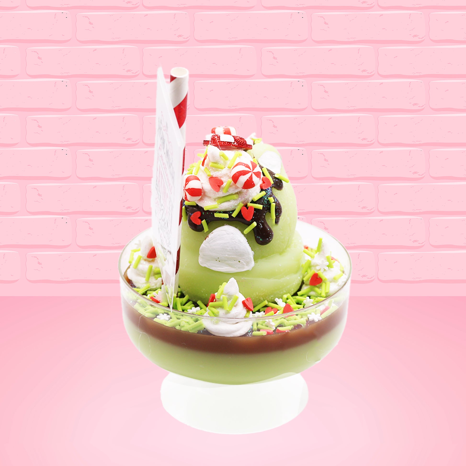 Cute kawaii ice cream Emotional Grinch Fudge sundae which has a sign and green white and red heart sprinkles inside of the dish as well as swirls of whipped cream. The bunny has chocolate glaze, peppermint candies and a swirl of whipped cream on it’s head. This view is from the back.