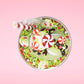 Cute kawaii ice cream Emotional Grinch Fudge sundae which has a sign and green white and red heart sprinkles inside of the dish as well as swirls of whipped cream. The bunny has chocolate glaze, peppermint candies and a swirl of whipped cream on it’s head. This view is from the top.