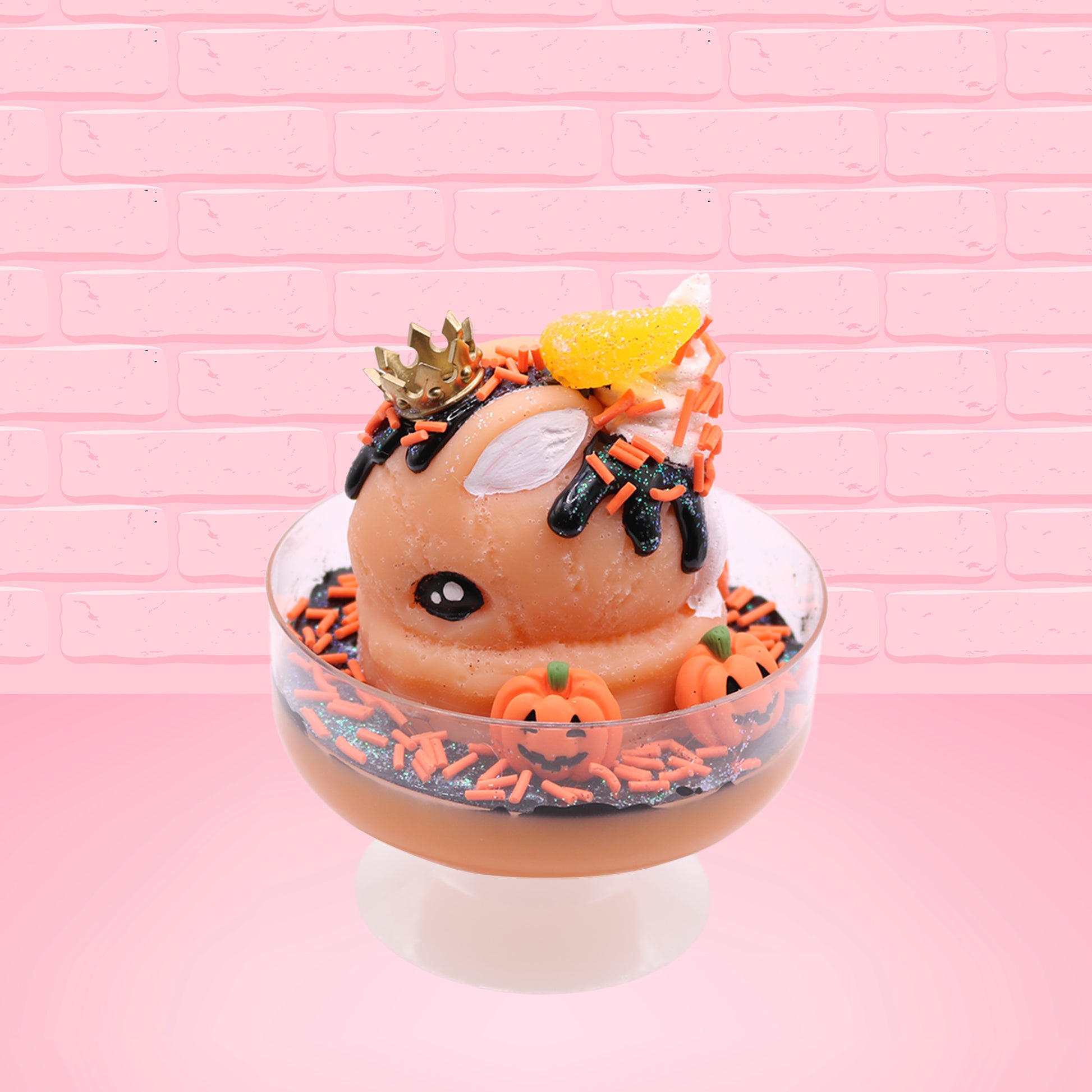 Cute kawaii Pumpkin King Bunny orange ice cream sundae with black glaze, orange sprinkles and pumpkin faces in the dish.  The bunny has a golden crown and orange slice on top next to the swirl of whipped cream. This view is from the side.