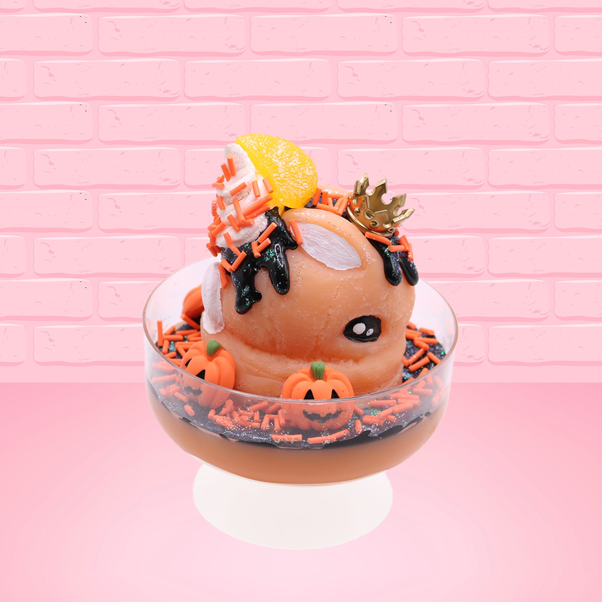 Cute kawaii Pumpkin King Bunny orange ice cream sundae with black glaze, orange sprinkles and pumpkin faces in the dish.  The bunny has a golden crown and orange slice on top next to the swirl of whipped cream. This view is from the other side.
