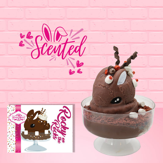 Cute kawaii bunny rocky on the rocks ice cream brown sundae with marshmallows and brown bits in the dish. The marshmallows on the top of the ice cream have red berries as well as two swirls of solid chocolate that stick up and a swirl of whipped cream is on the back. There are two chocolate balls and one red ball on between the ears of the ice cream bunny. The front of the character card and the word scented is shown.