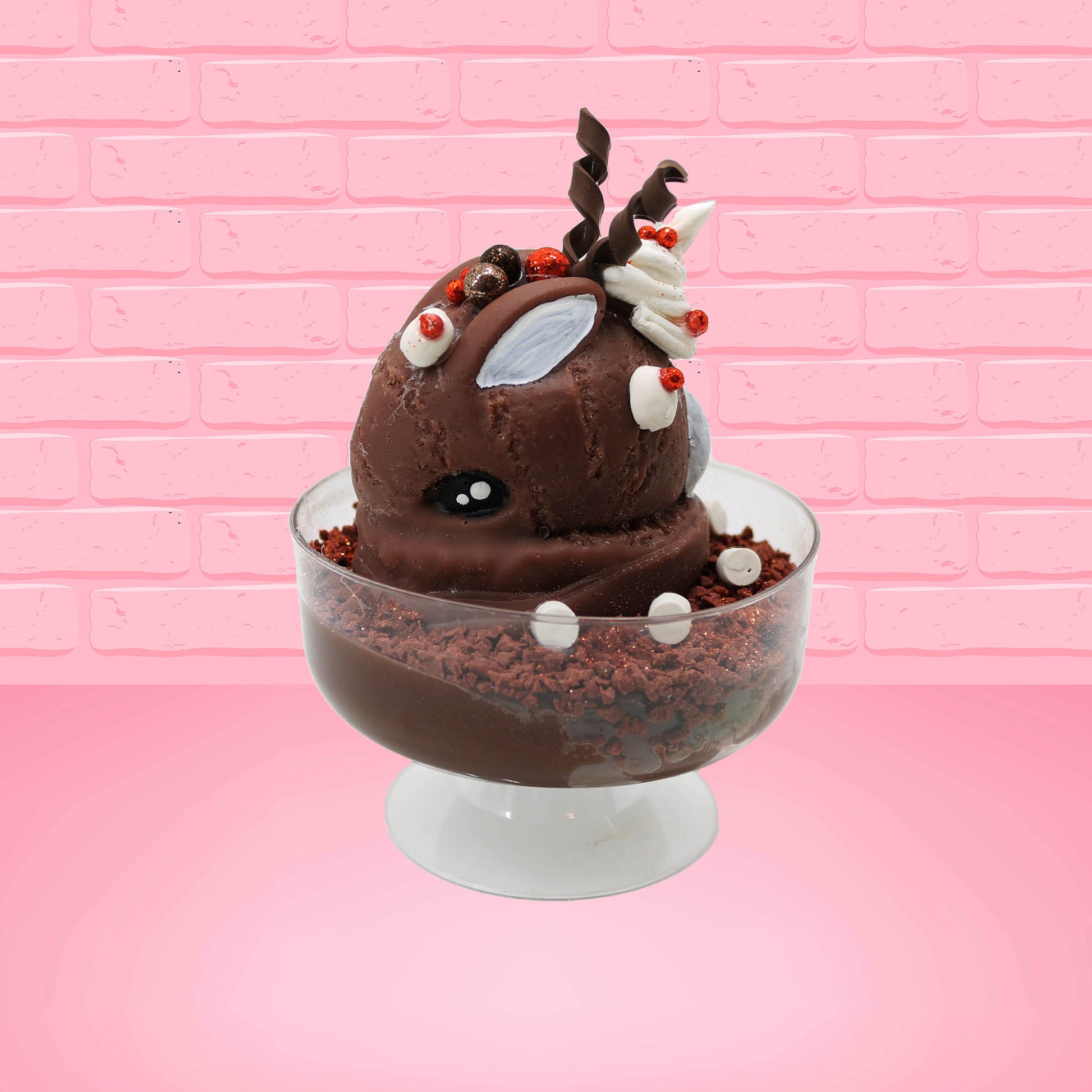 Cute kawaii bunny rocky on the rocks ice cream brown sundae with marshmallows and brown bits in the dish. The marshmallows on the top of the ice cream have red berries as well as two swirls of solid chocolate that stick up and a swirl of whipped cream is on the back. There are two chocolate balls and one red ball on between the ears of the ice cream bunny. This view is from the side.
