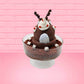 Cute kawaii bunny rocky on the rocks ice cream brown sundae with marshmallows and brown bits in the dish. The marshmallows on the top of the ice cream have red berries as well as two swirls of solid chocolate that stick up and a swirl of whipped cream is on the back. There are two chocolate balls and one red ball on between the ears of the ice cream bunny. This view is from the back.