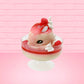 Cute kawaii bunny strawberry cheesecake white ice cream with red glaze, strawberries, and whipped cream swirls in the dish. The bunny has a strawberry on top between the ears, with a swirl of whipped cream on the back. This view is from the front at an angle.