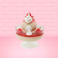 Cute kawaii bunny strawberry cheesecake white ice cream with red glaze, strawberries, and whipped cream swirls in the dish. The bunny has a strawberry on top between the ears, with a swirl of whipped cream on the back. This view is from the back.