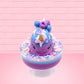 Cute kawaii unicorn bunny ice cream Sugar Berry Twilight sundae with whipped cream swirls, pink, blue, and white sprinkles in the dish. The unicorn bunny is purple and blue on top with blue berries and light purple raspberries, with a whipped cream swirl. This view is from the front.