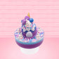 Cute kawaii unicorn bunny ice cream Sugar Berry Twilight sundae with whipped cream swirls, pink, blue, and white sprinkles in the dish. The unicorn bunny is purple and blue on top with blue berries and light purple raspberries, with a whipped cream swirl. This view is from the back.