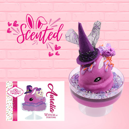 Cute kawaii witch bunny pink ice cream sundae with purple tall hat with translucent black layer covered with pink and purple crystals in the dish. The bunny has light purple raspberries near the swirl of whipped cream, and orange sprinkles in the light purple drippings. The character card front is shown, and the word scented.