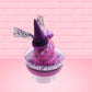 Cute kawaii witch bunny pink ice cream sundae with purple tall hat with translucent black layer covered with pink and purple crystals in the dish. The bunny has light purple raspberries near the swirl of whipped cream, and orange sprinkles in the light purple drippings. This view is from the front.