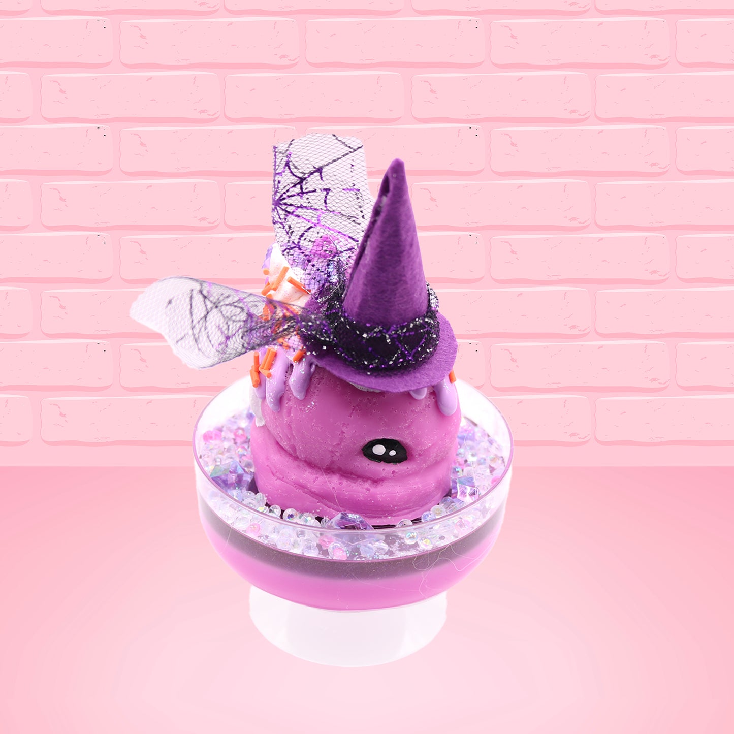 Cute kawaii witch bunny pink ice cream sundae with purple tall hat with translucent black layer covered with pink and purple crystals in the dish. The bunny has light purple raspberries near the swirl of whipped cream, and orange sprinkles in the light purple drippings. This view is from the front at an angle.