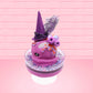 Cute kawaii witch bunny pink ice cream sundae with purple tall hat with translucent black layer covered with pink and purple crystals in the dish. The bunny has light purple raspberries near the swirl of whipped cream, and orange sprinkles in the light purple drippings. This view is from the side.
