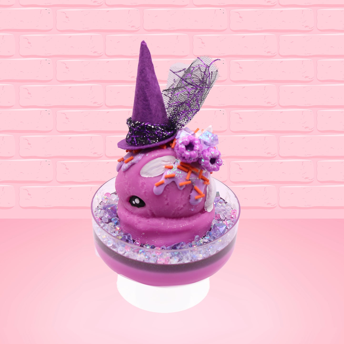 Cute kawaii witch bunny pink ice cream sundae with purple tall hat with translucent black layer covered with pink and purple crystals in the dish. The bunny has light purple raspberries near the swirl of whipped cream, and orange sprinkles in the light purple drippings. This view is from the side.