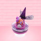 Cute kawaii witch bunny pink ice cream sundae with purple tall hat with translucent black layer covered with pink and purple crystals in the dish. The bunny has light purple raspberries near the swirl of whipped cream, and orange sprinkles in the light purple drippings. This view is from the back.