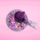 Cute kawaii witch bunny pink ice cream sundae with purple tall hat with translucent black layer covered with pink and purple crystals in the dish. The bunny has light purple raspberries near the swirl of whipped cream, and orange sprinkles in the light purple drippings. This view is from the top.