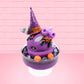 Cute kawaii witch bunny Blair purple ice cream with purple crystals and pumpkins on top of a layer of glittery black translucent layer. The bunny has a purple witch’s hat, with purple raspberries and a whipped cream swirl on top, with black glittery glaze and orange sprinkles. This view is from the other side.