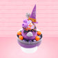 Cute kawaii witch bunny Blair purple ice cream with purple crystals and pumpkins on top of a layer of glittery black translucent layer. The bunny has a purple witch’s hat, with purple raspberries and a whipped cream swirl on top, with black glittery glaze and orange sprinkles. This view is from the back.