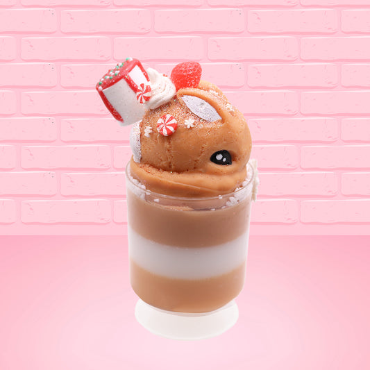Cute kawaii bunny Gingerbread Mousse tall ice cream sundae that has white crumbles, a gingerbread man with a santa hat, and a marshmallow with glittery red glaze in the dish.  The bunny has peppermint candies, a strawberry, and a swirl of whipped cream on top and a snowflake on the front. This view is from the side.