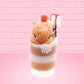Cute kawaii bunny Gingerbread Mousse tall ice cream sundae that has white crumbles, a gingerbread man with a santa hat, and a marshmallow with glittery red glaze in the dish.  The bunny has peppermint candies, a strawberry, and a swirl of whipped cream on top and a snowflake on the front. This view is from the front.