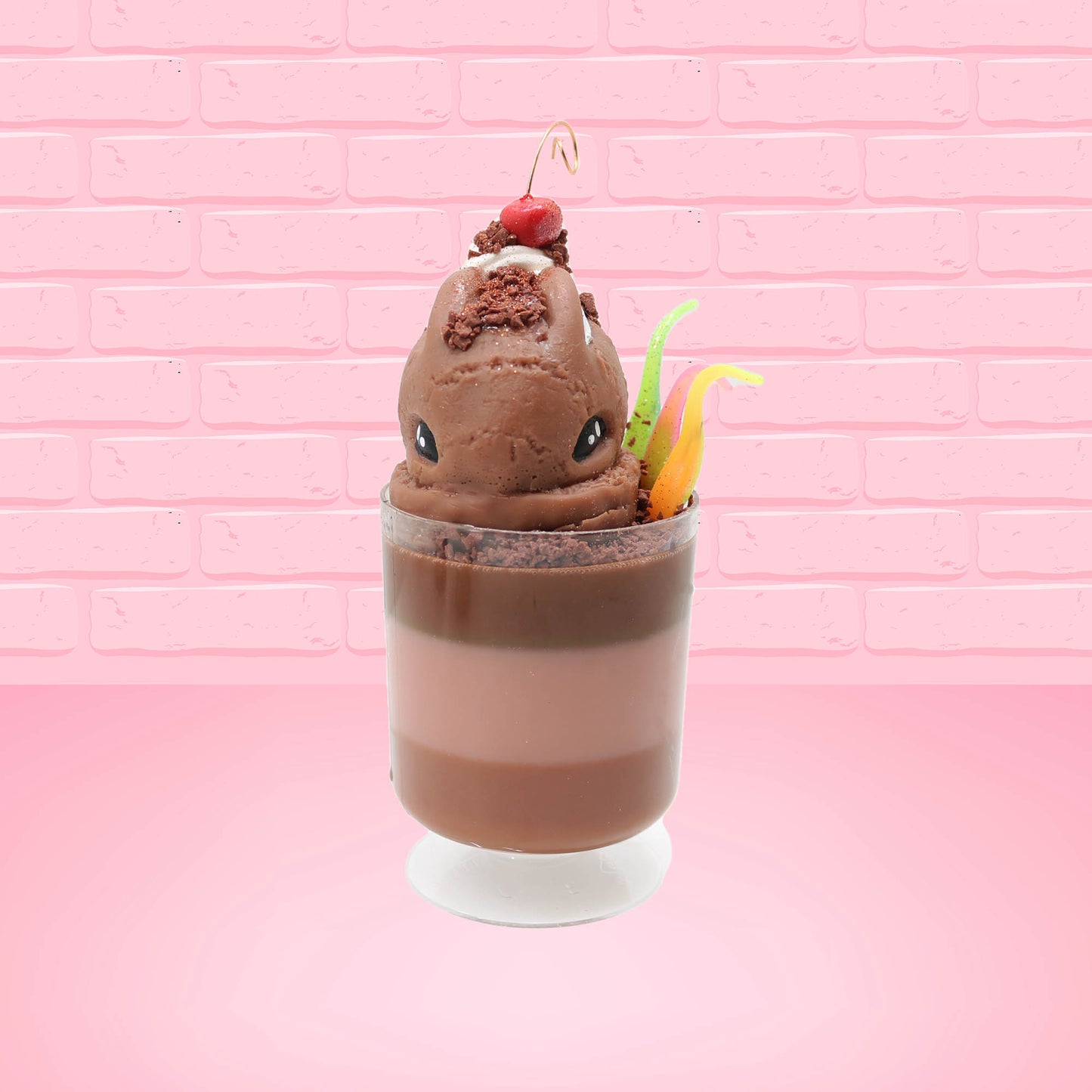 Cute kawaii bunny tall brown and light brown ice cream sundae with brown chocolate crumbles over the bunny, with a cherry on top of the whipped cream swirl.  Three bendy gummy worms are in the dish on the side. This view is from the front.