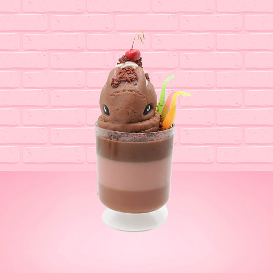 Cute kawaii bunny tall brown and light brown ice cream sundae with brown chocolate crumbles over the bunny, with a cherry on top of the whipped cream swirl.  Three bendy gummy worms are in the dish on the side. This view is from the front.