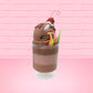 Cute kawaii bunny tall brown and light brown ice cream sundae with brown chocolate crumbles over the bunny, with a cherry on top of the whipped cream swirl.  Three bendy gummy worms are in the dish on the side. This view is from the front at an angle.