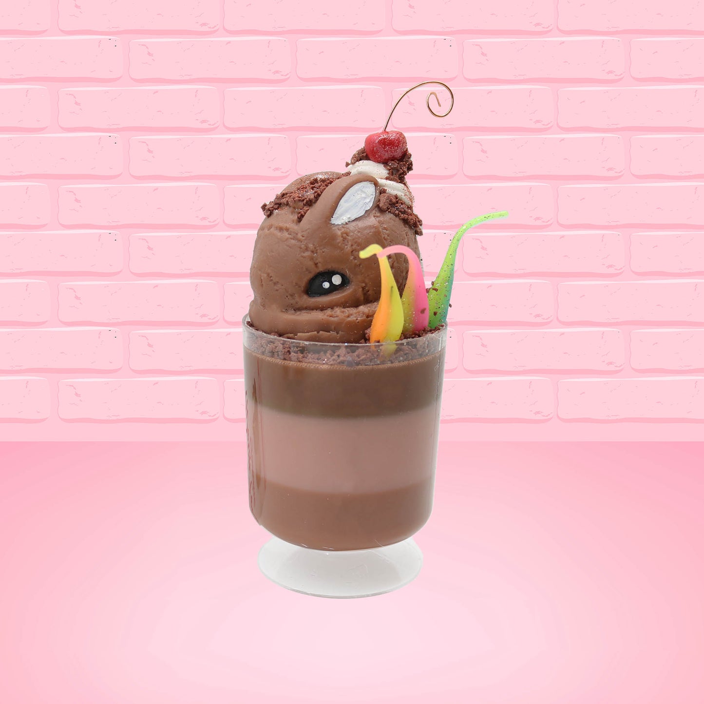 Cute kawaii bunny tall brown and light brown ice cream sundae with brown chocolate crumbles over the bunny, with a cherry on top of the whipped cream swirl.  Three bendy gummy worms are in the dish on the side. This view is from the front at an angle.