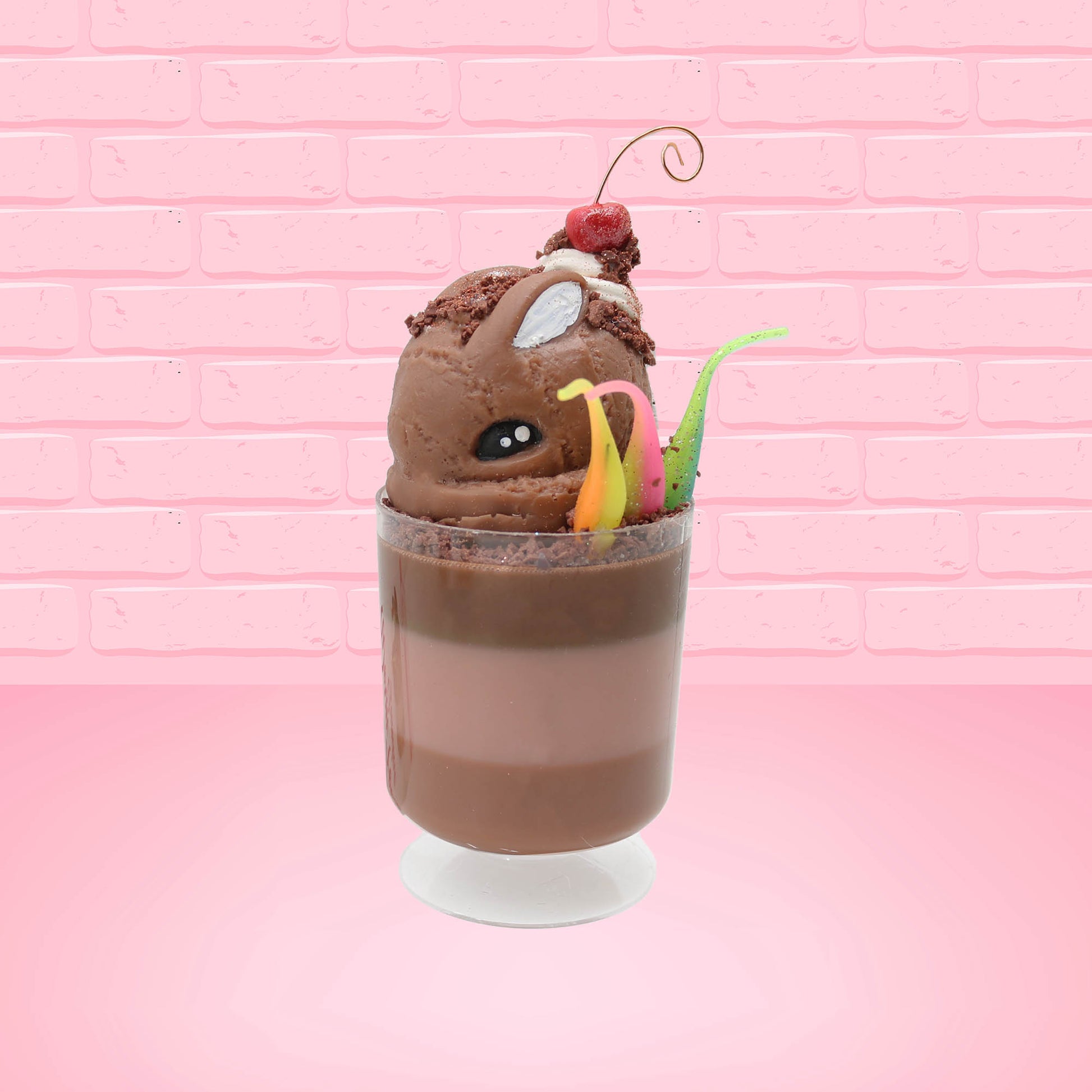 Cute kawaii bunny tall brown and light brown ice cream sundae with brown chocolate crumbles over the bunny, with a cherry on top of the whipped cream swirl.  Three bendy gummy worms are in the dish on the side. This view is from the front at an angle.