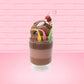 Cute kawaii bunny tall brown and light brown ice cream sundae with brown chocolate crumbles over the bunny, with a cherry on top of the whipped cream swirl.  Three bendy gummy worms are in the dish on the side. This view is from the side.